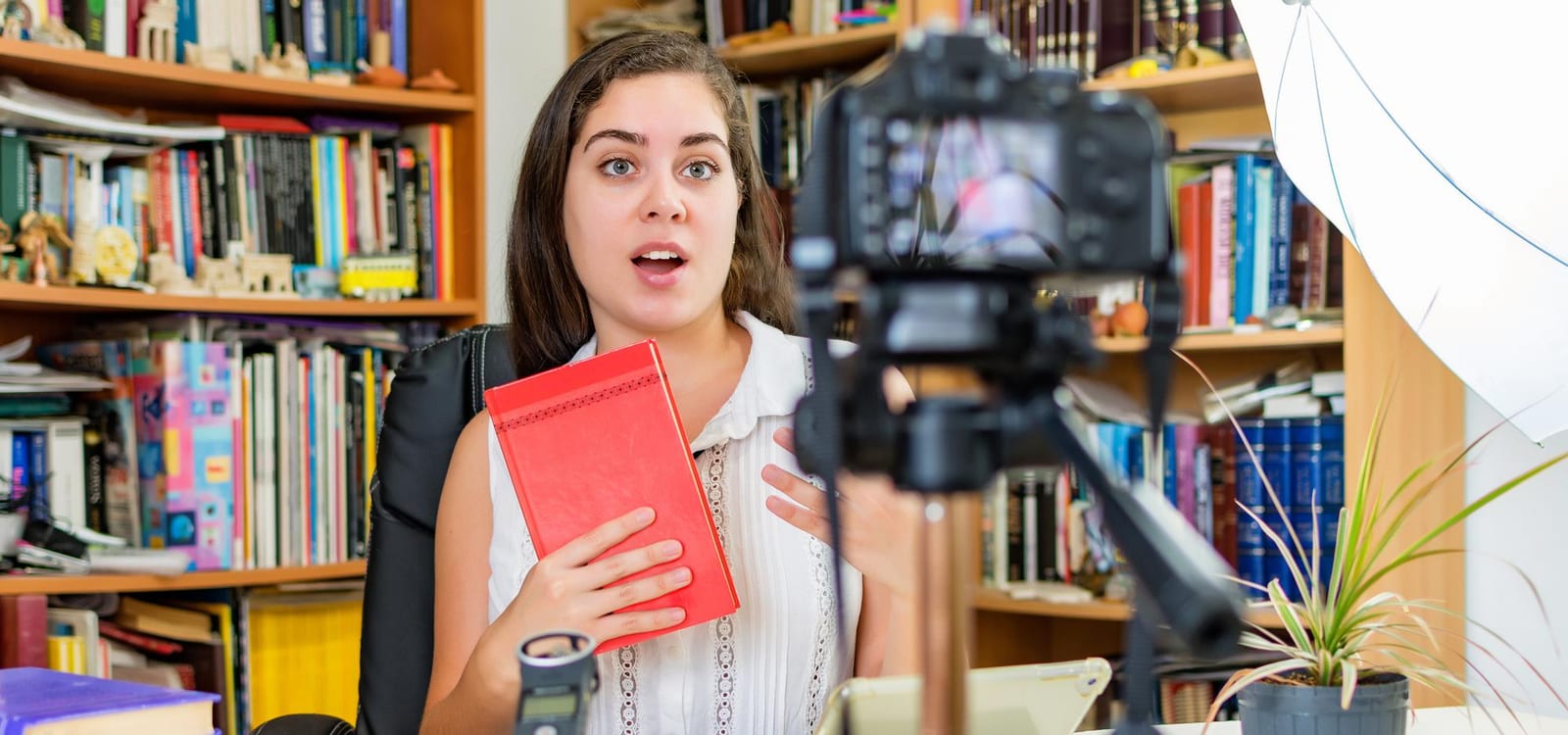 Mulher segura um livro fechado diante de estantes cheias de livros, em uma sala de leitura.